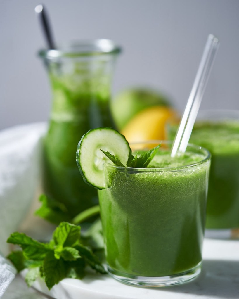 Green juice surrounded by fresh spinach, topped with cucumber and lemon